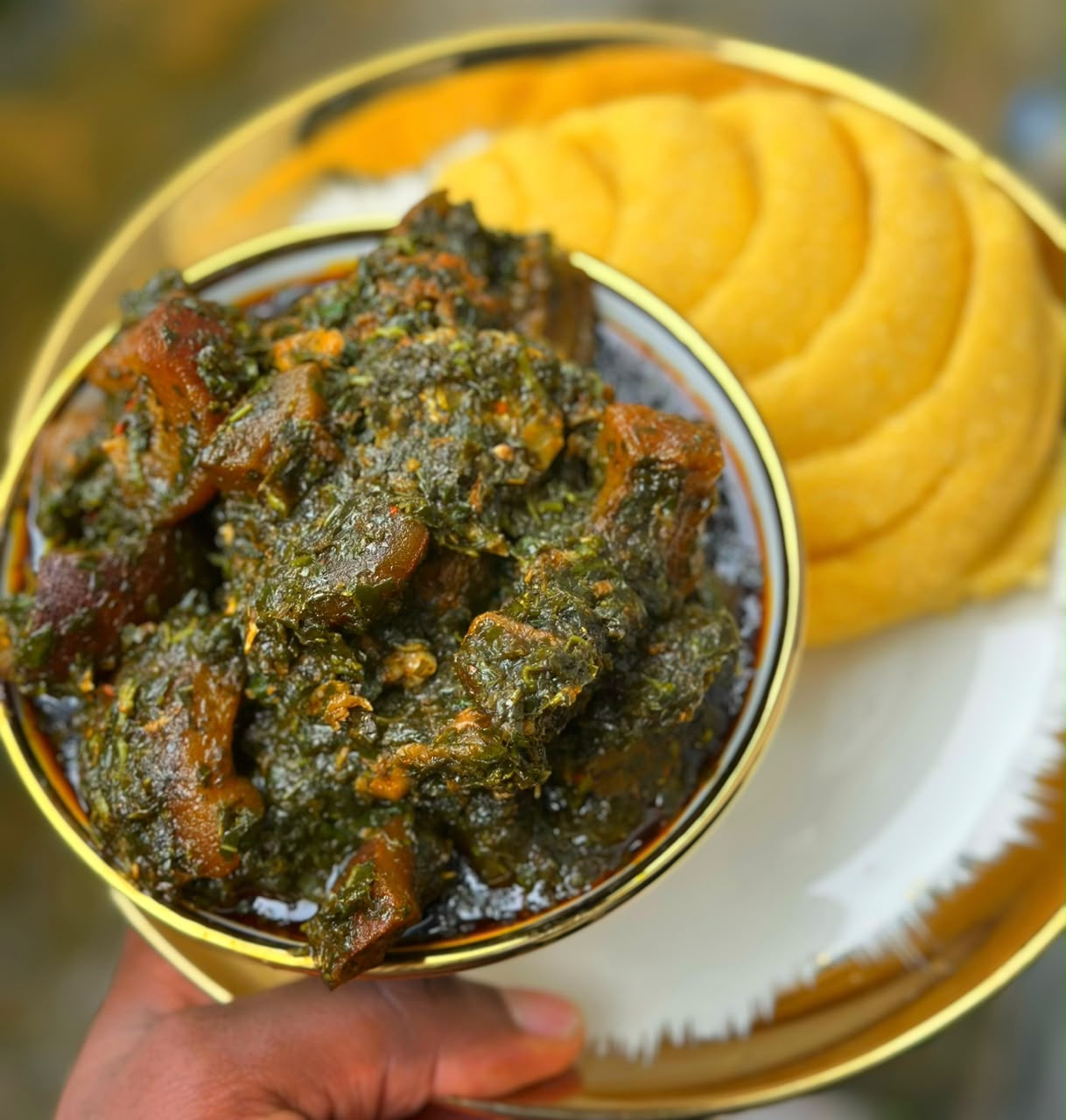 A bowl of rich Akwa Ibom vegetable soup in a gold-rimmed bowl served with yellow eba.